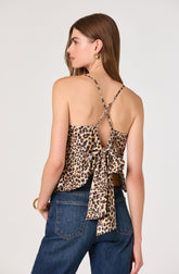 A woman is wearing a sleeveless top with thin, leopard-print spaghetti straps and a large bow tied at the back. The top features a leopard print pattern with shades of tan, brown, and black. She pairs it with high-waisted dark blue jeans.